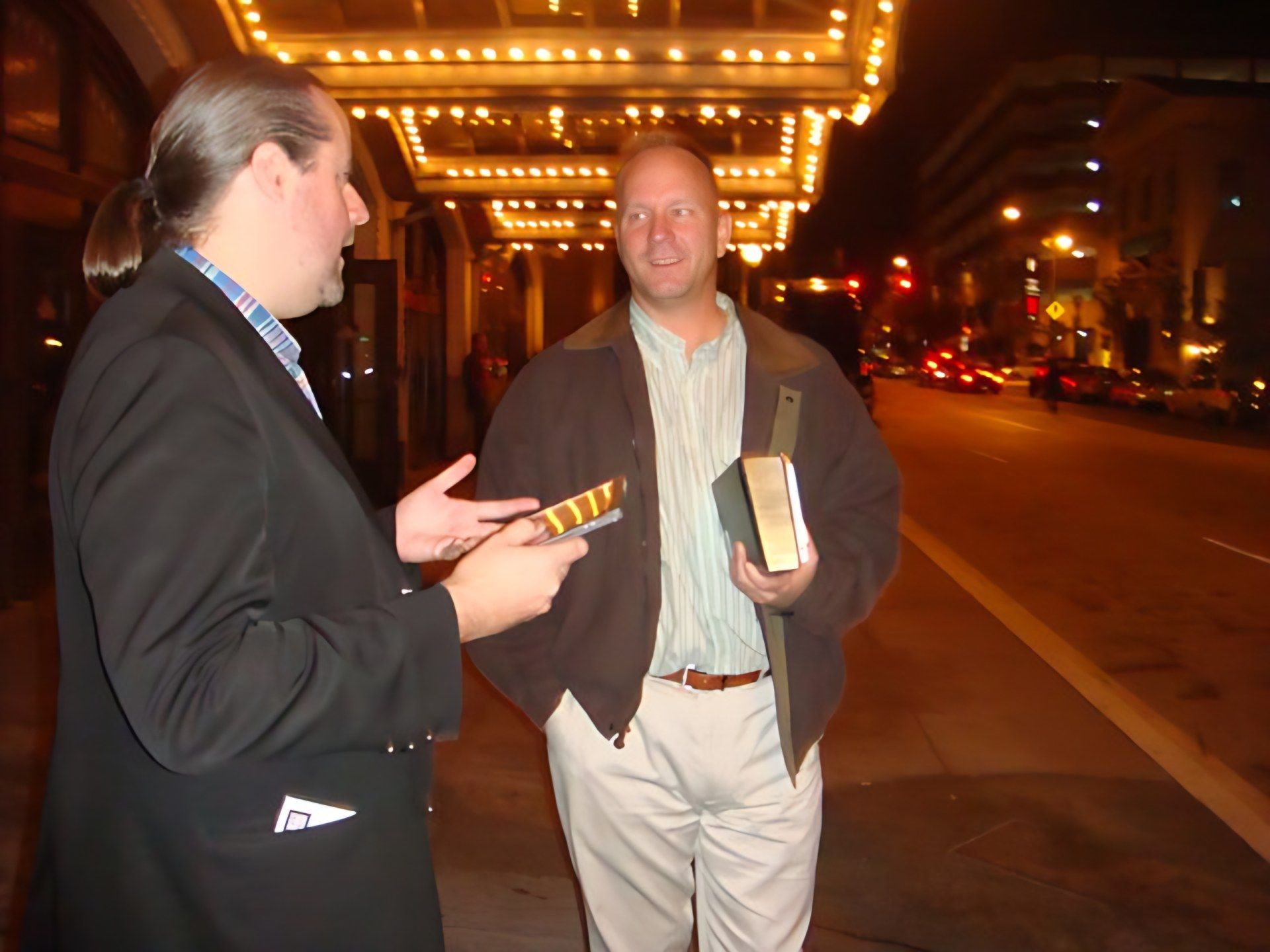 Two men talking on the sidewalk at night under a theater marquee lined with light bulbs, one holding a book