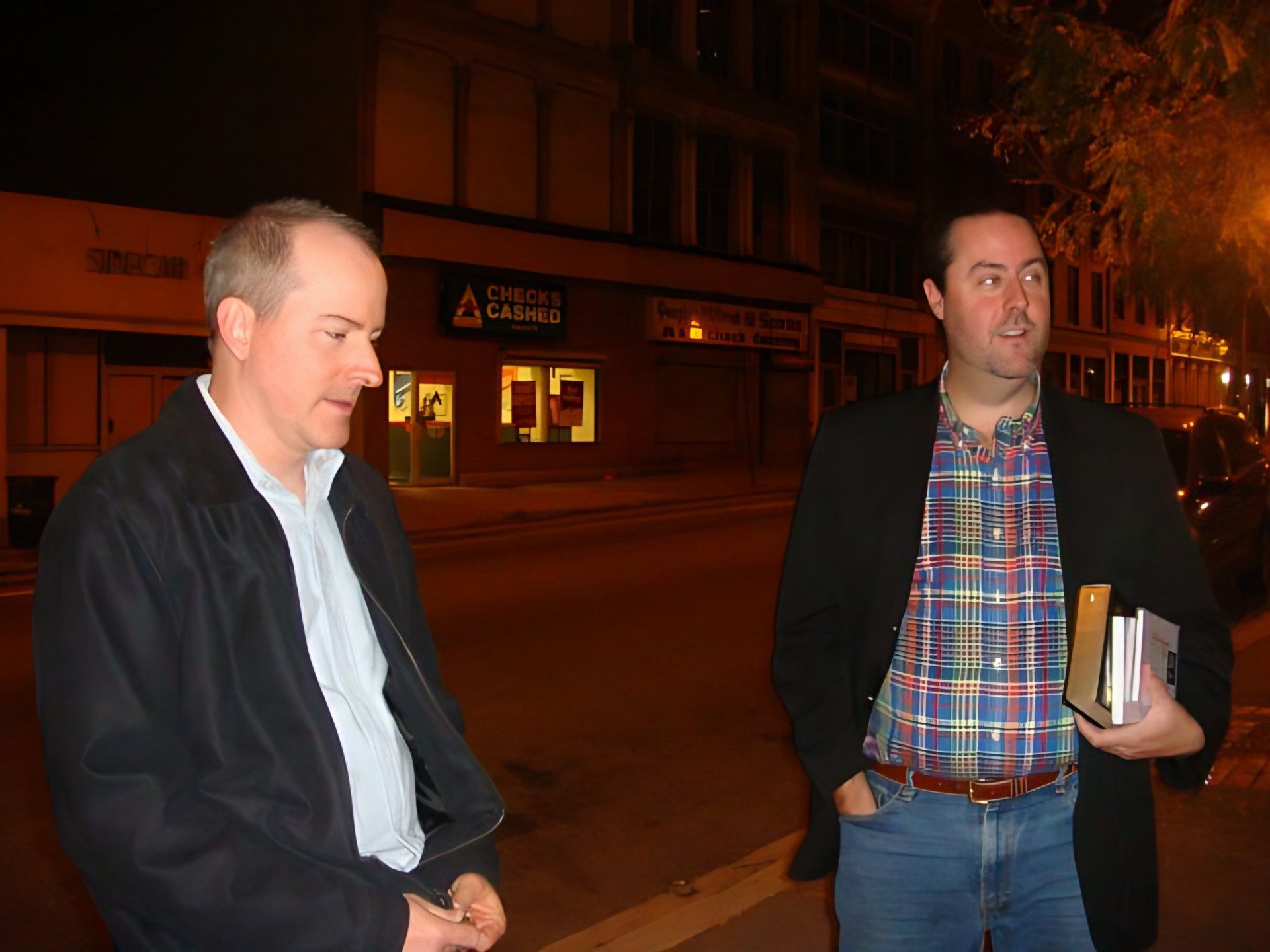 Sean Fenlon, in a plaid shirt and dark sports coat and holding books, talks with a friend on a Baltimore street at night
