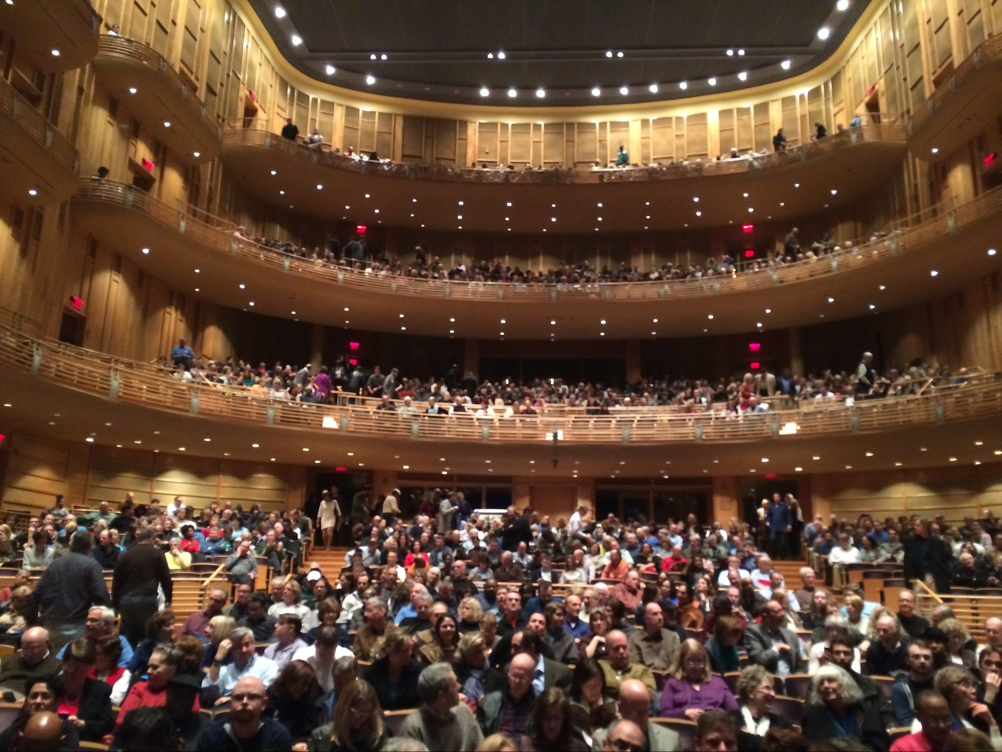 A packed concert hall seen from near the stage, looking back over the orchestra seats to three curved balcony tiers under a ceiling of small lights