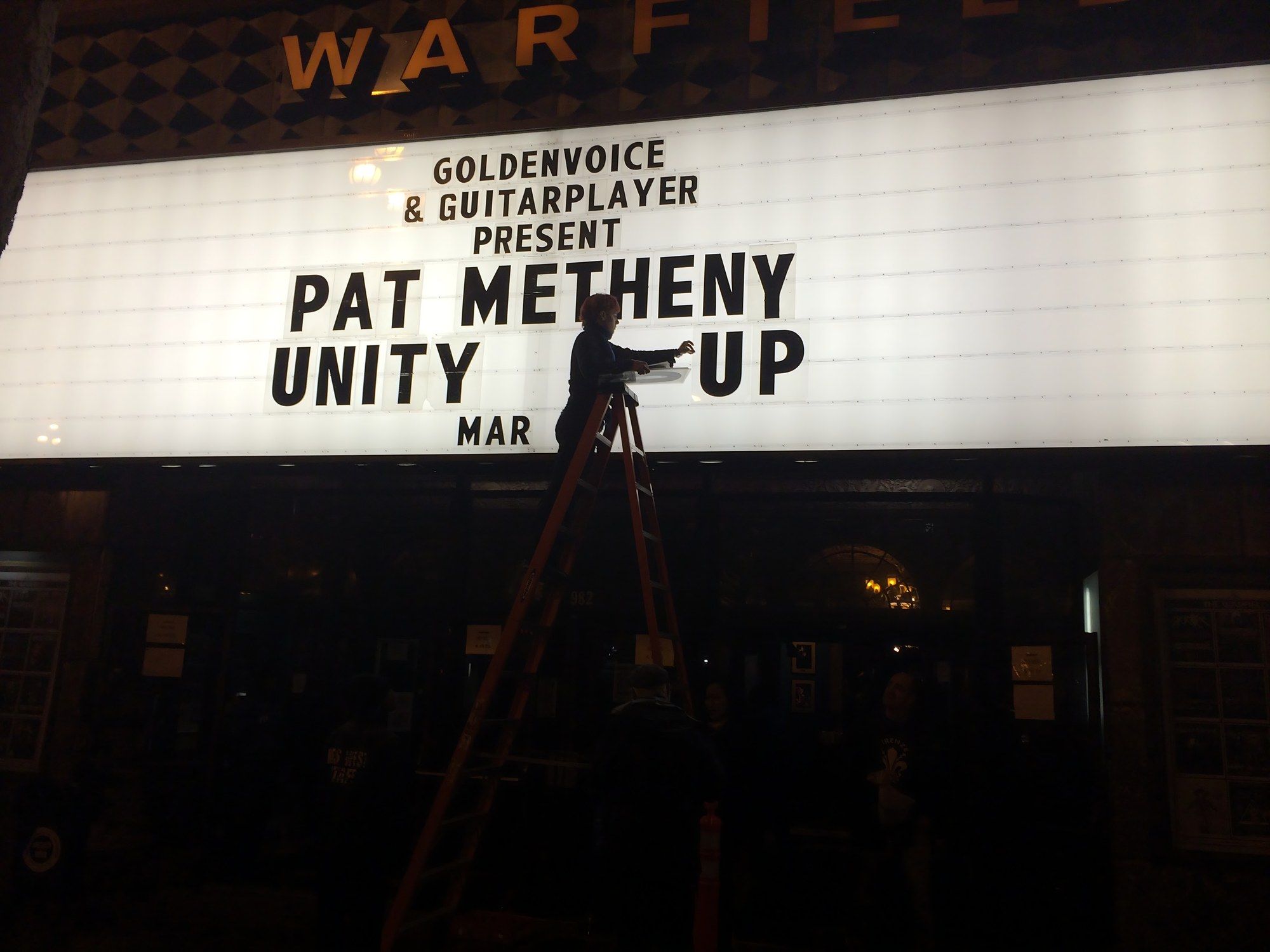 Close view of The Warfield marquee reading “Goldenvoice and Guitar Player present Pat Metheny Unity Group,” with a worker on a ladder changing the letters